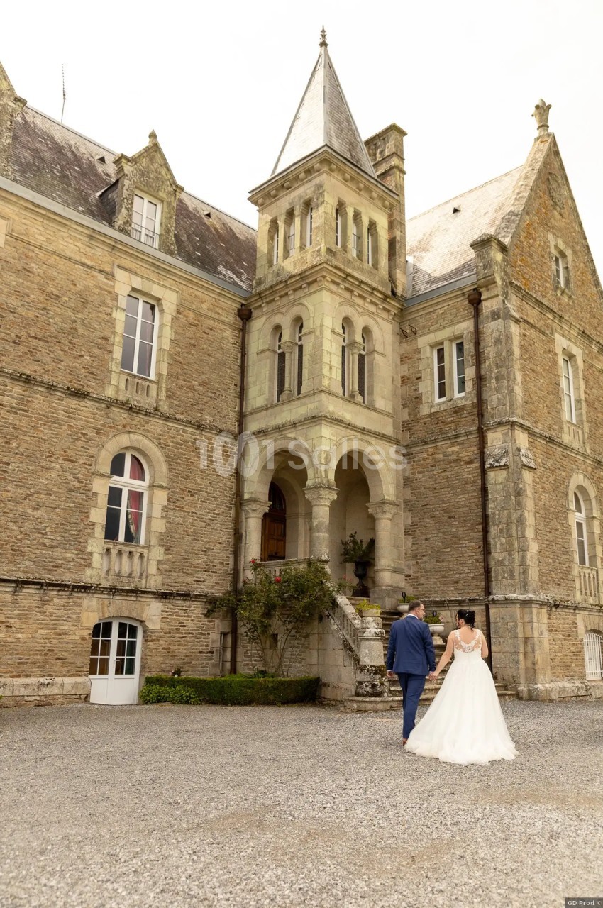 Un couple en tenue de mariage marche vers un château en pierre sous un ciel clair.