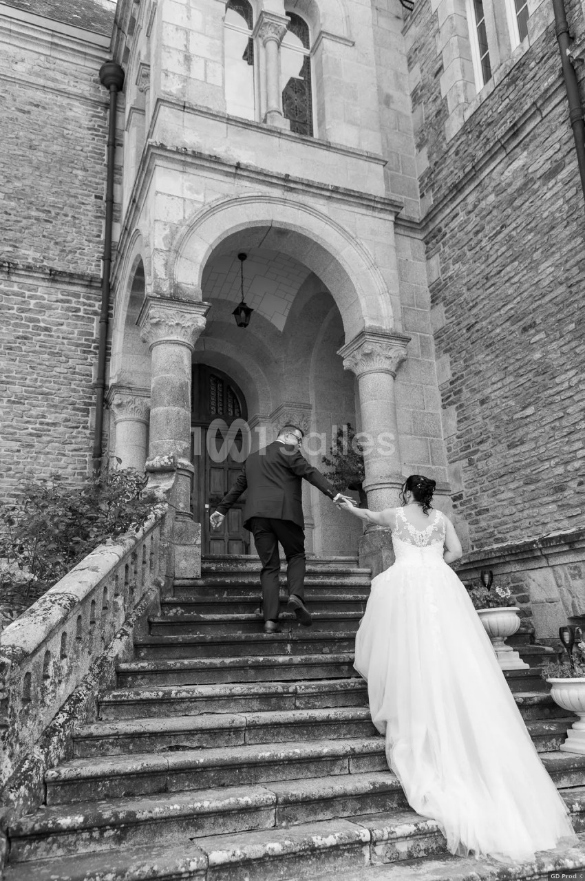 Un couple monte les marches d'un bâtiment ancien en pierre, la mariée en robe blanche tenant la main du marié.