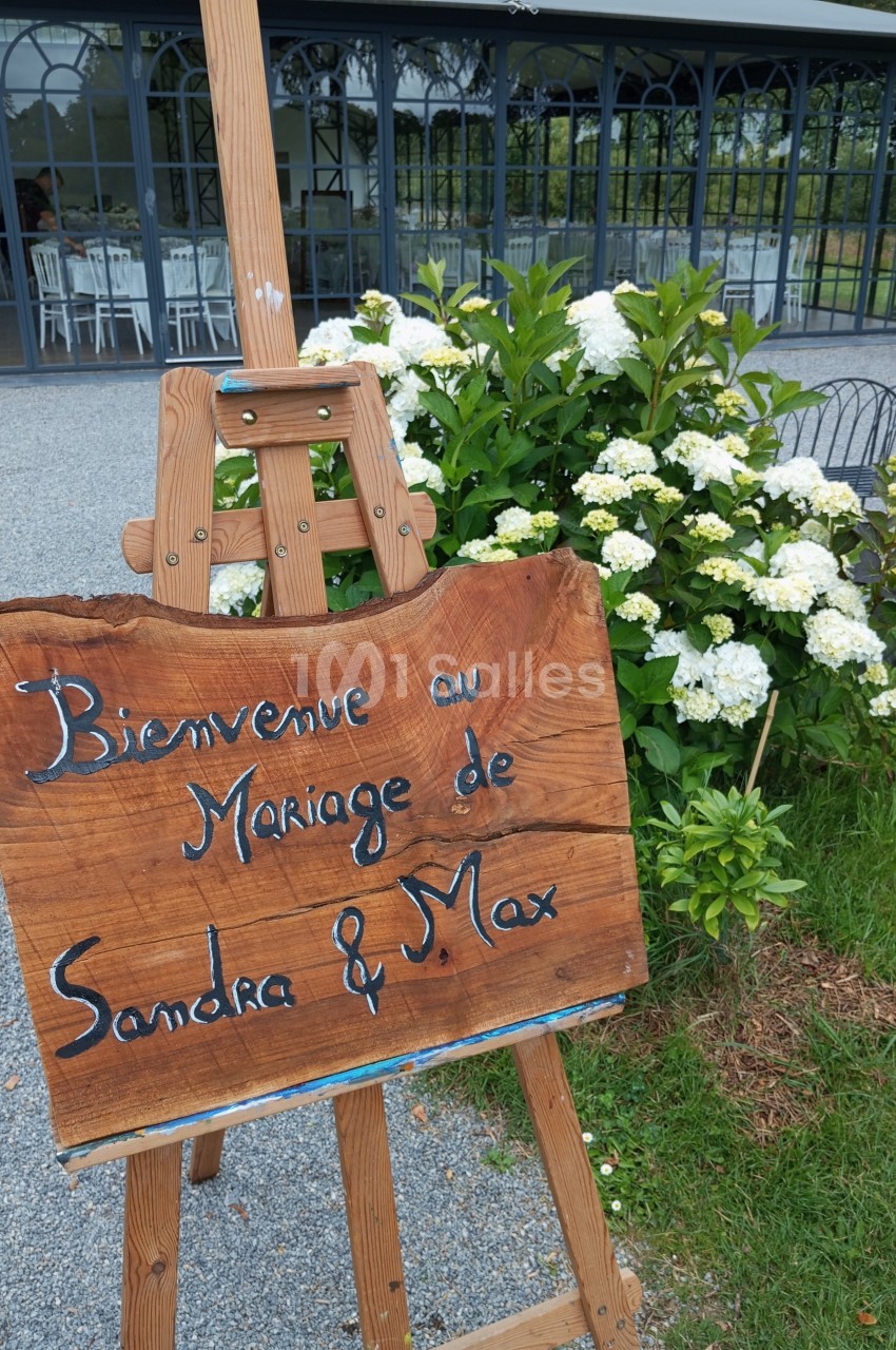 Panneau en bois sur chevalet indiquant ’Bienvenue au mariage de Sandra & Max’, devant un jardin et une salle vitrée.