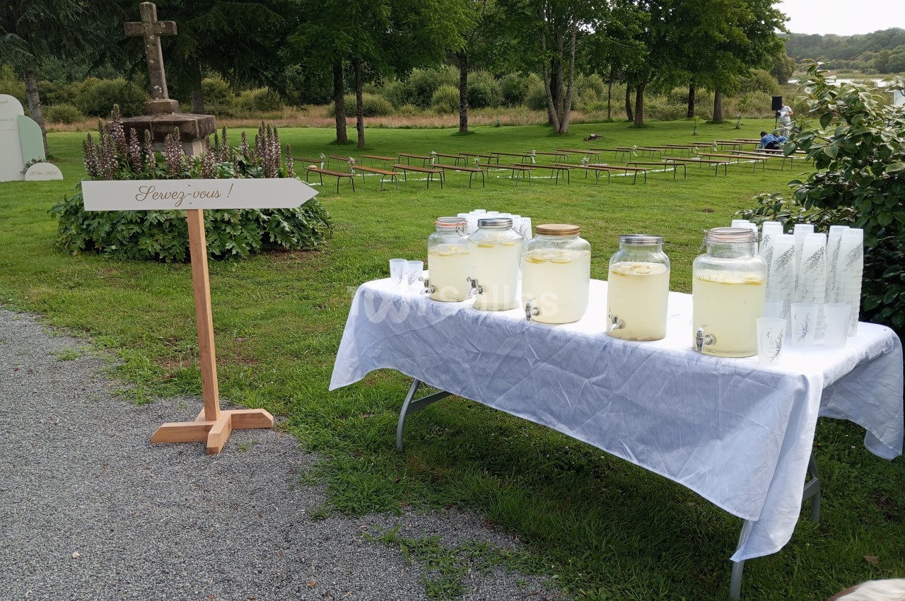 Table avec des bocaux de limonade et des verres, installée en extérieur près d'une flèche indiquant ’Servez-vous’.