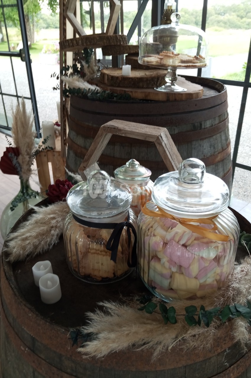 Bonbonnières en verre remplies de friandises colorées, posées sur des tonneaux en bois dans un décor champêtre.