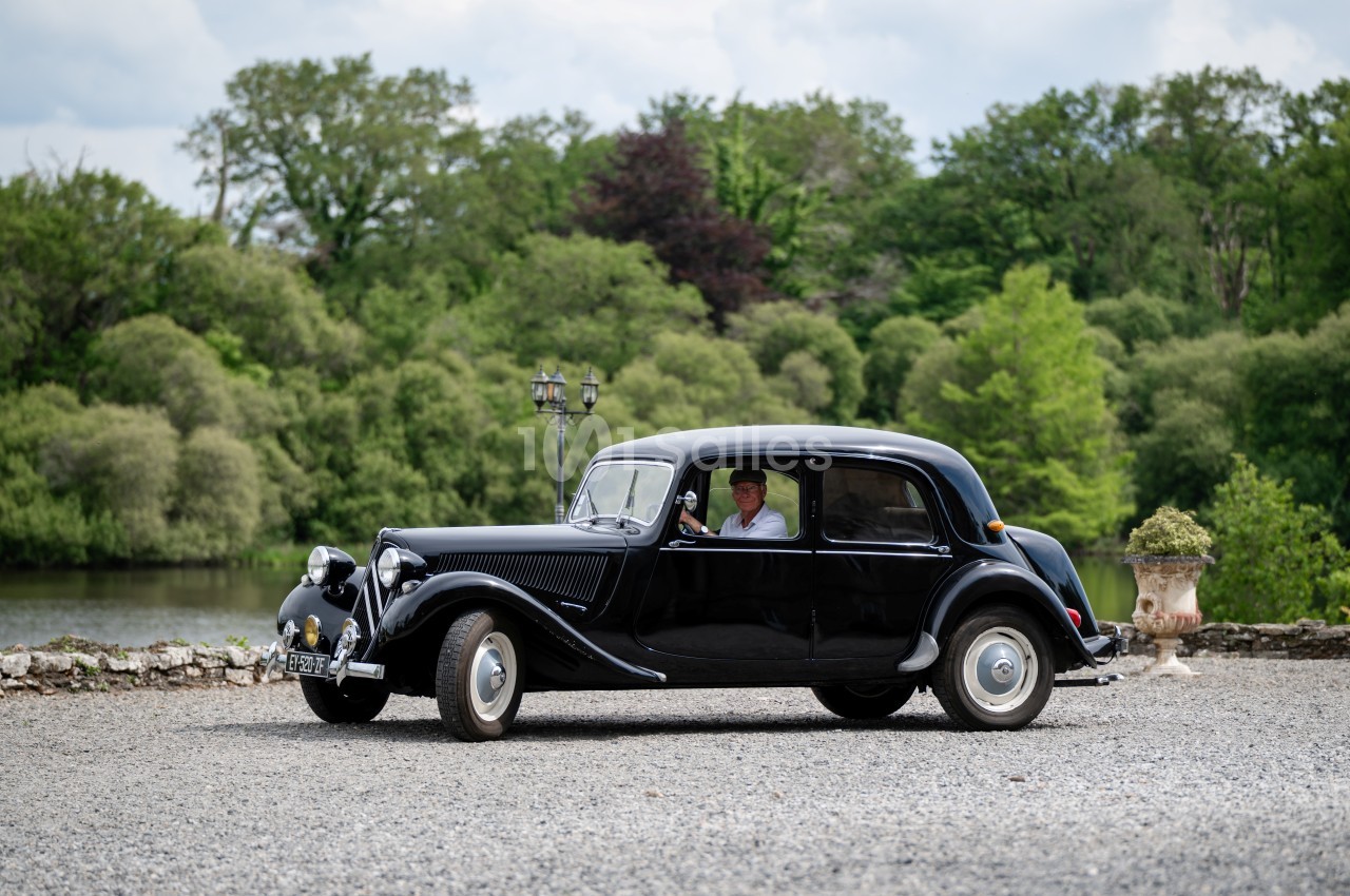 Voiture ancienne noire stationnée sur un chemin gravillonné, avec un conducteur visible, entourée de verdure.
