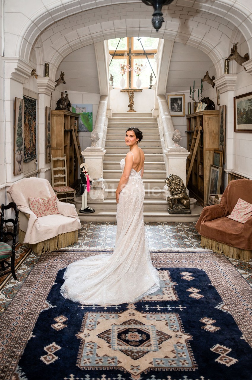 Femme en robe de mariée dans une pièce élégante avec tapis, fauteuils et escalier menant à une fenêtre lumineuse.