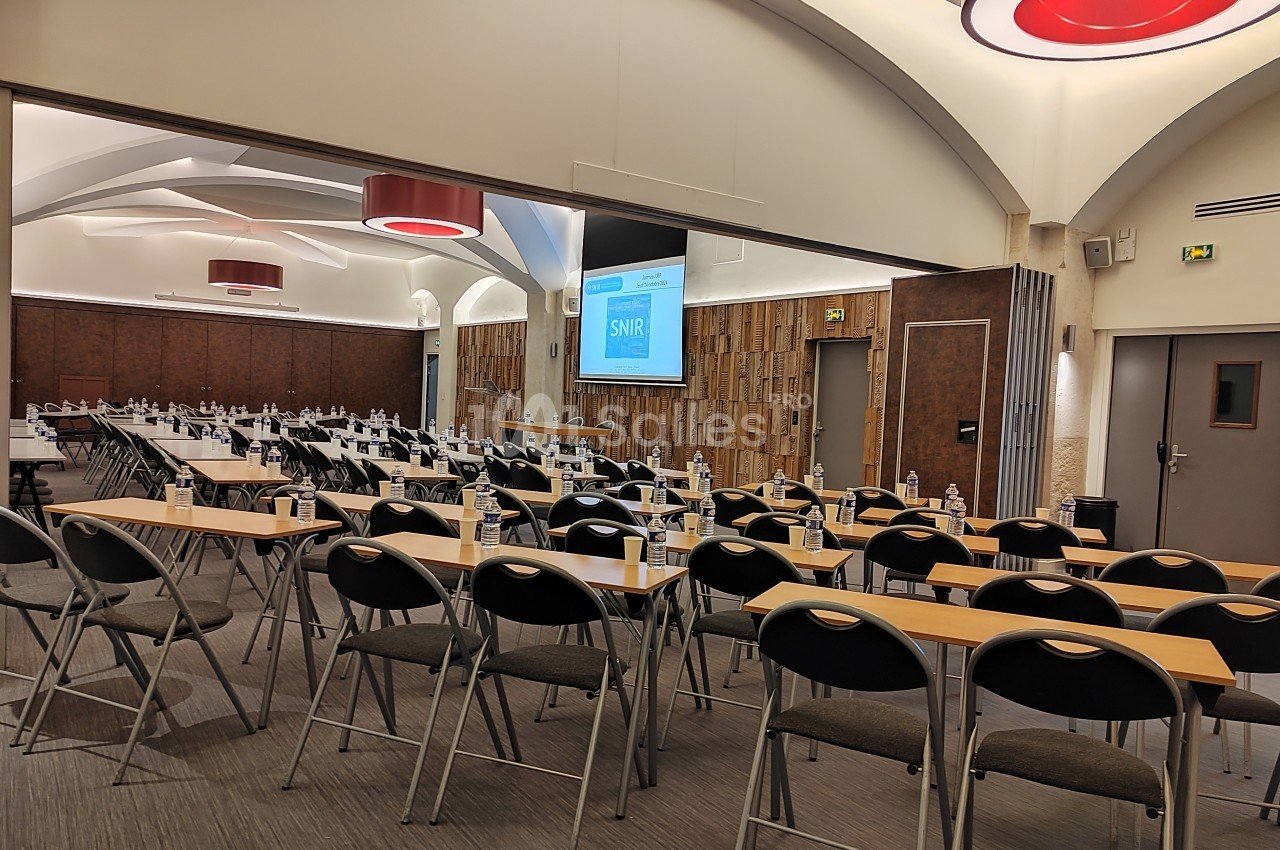 Salle de conférence avec des rangées de tables et chaises alignées, écran de projection allumé en arrière-plan.