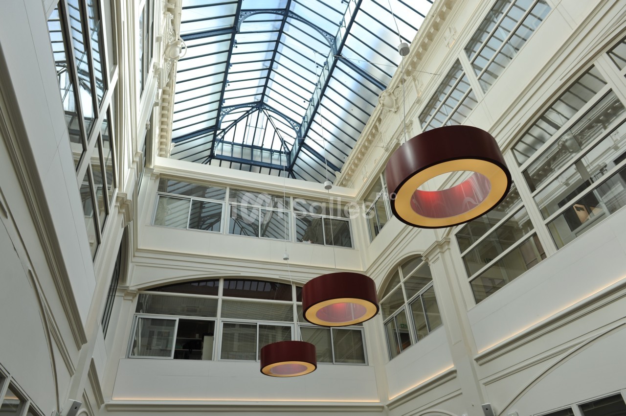 Vue intérieure d'une galerie lumineuse avec verrière, murs blancs et suspensions circulaires rouges.