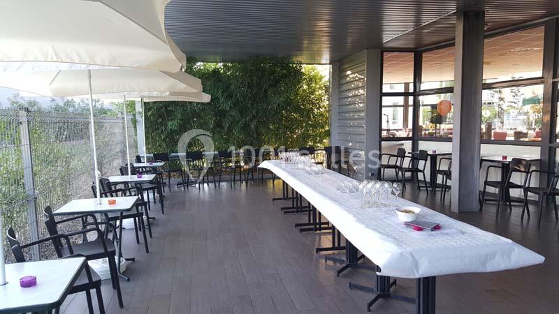 Terrasse couverte avec tables et chaises, grandes tables dressées avec verres et nappes blanches, végétation en arrière-plan.