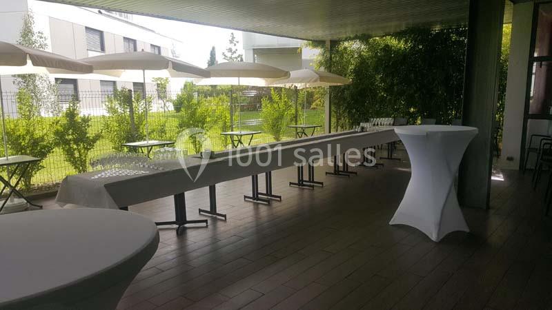 Terrasse couverte aménagée avec des tables hautes et une longue table dressée, entourée de verdure.