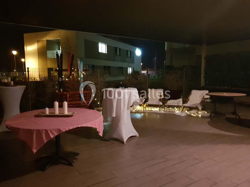 Terrasse éclairée de nuit avec tables décorées, chaises en bois et guirlandes lumineuses en arrière-plan.