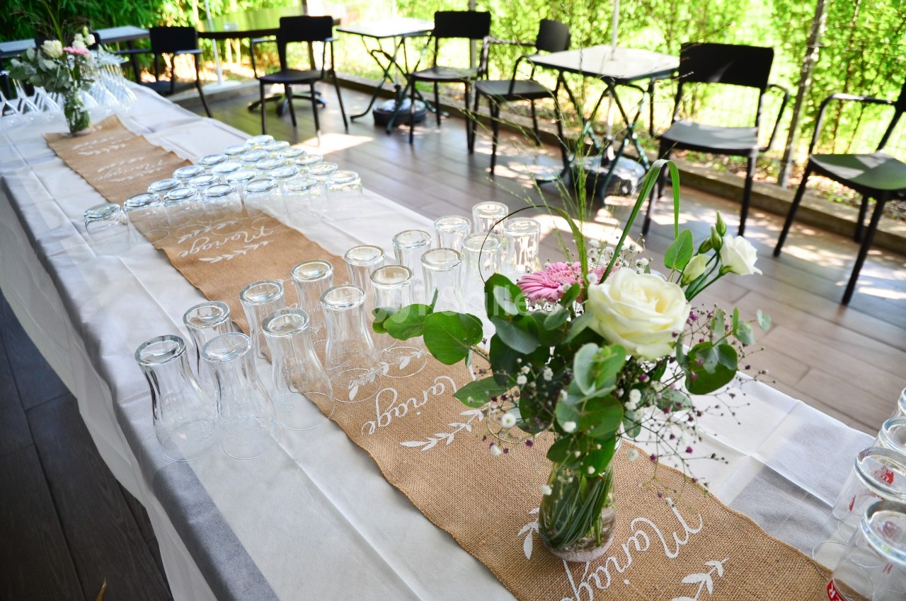 Table décorée avec nappes en jute, verres alignés et vase contenant des fleurs blanches et roses, en extérieur.