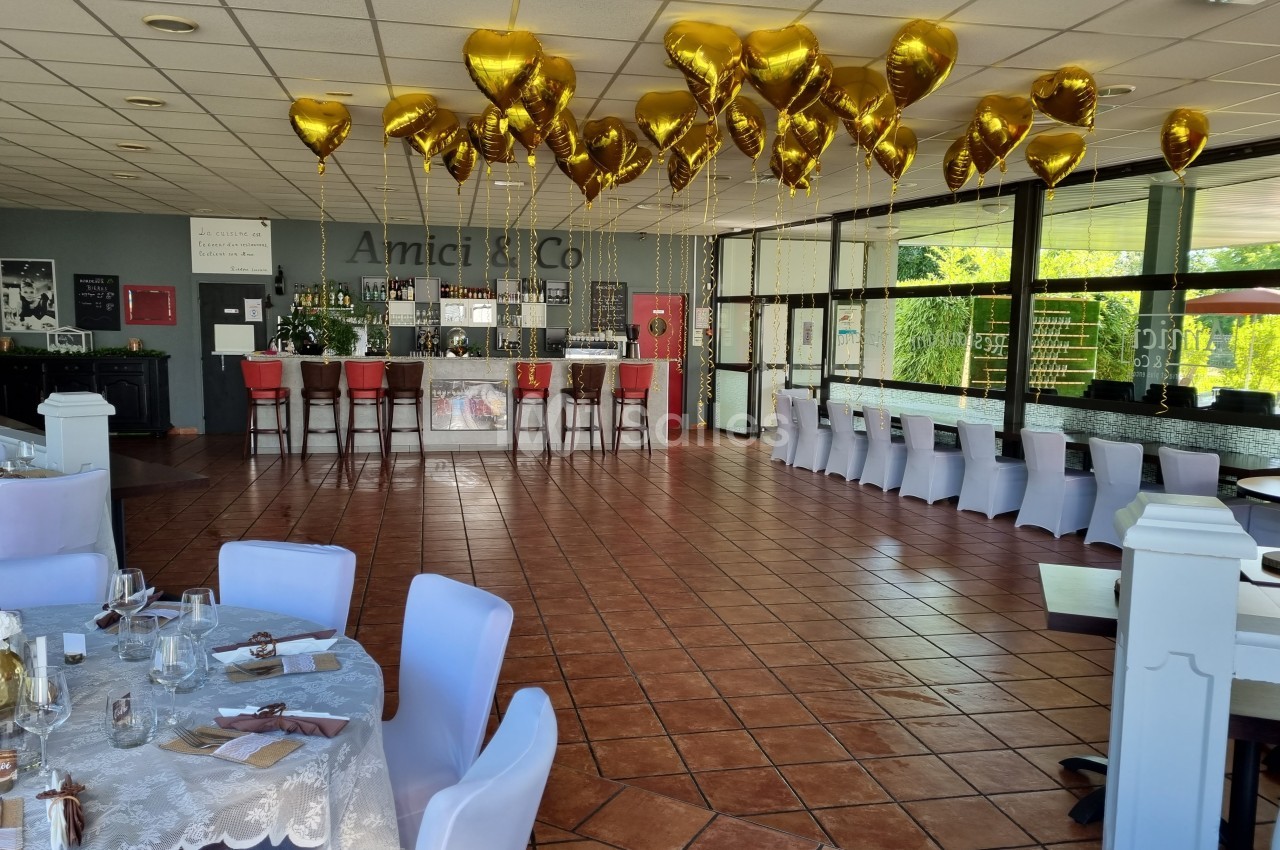 Salle de réception avec décoration de ballons dorés au plafond, tables dressées et bar en arrière-plan.