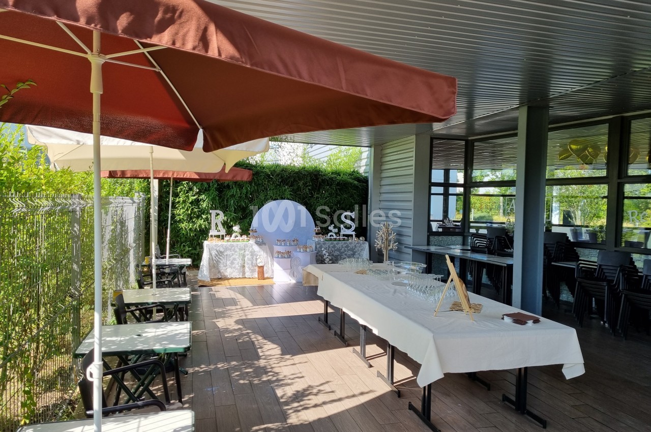Terrasse aménagée avec tables dressées pour un événement, décorations blanches et végétation en arrière-plan.