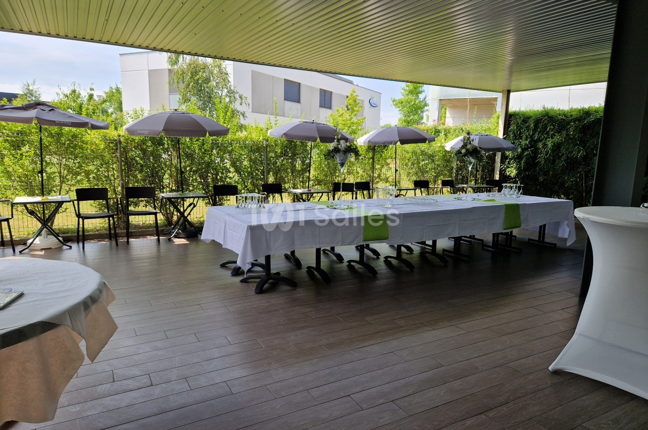 Terrasse couverte aménagée avec de longues tables dressées, chaises noires et parasols, entourée de verdure.