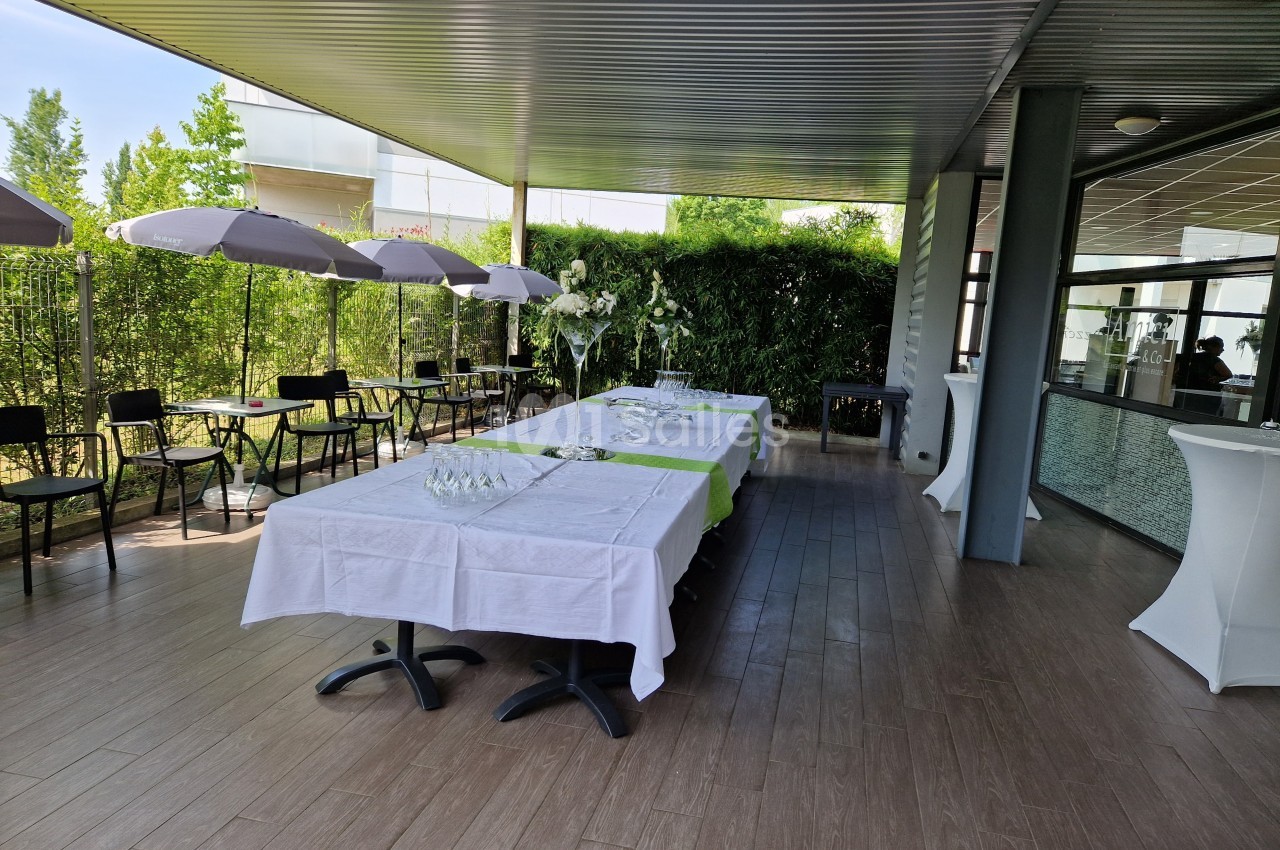 Terrasse aménagée avec tables dressées, nappes blanches, décorations florales et chaises sous des parasols.