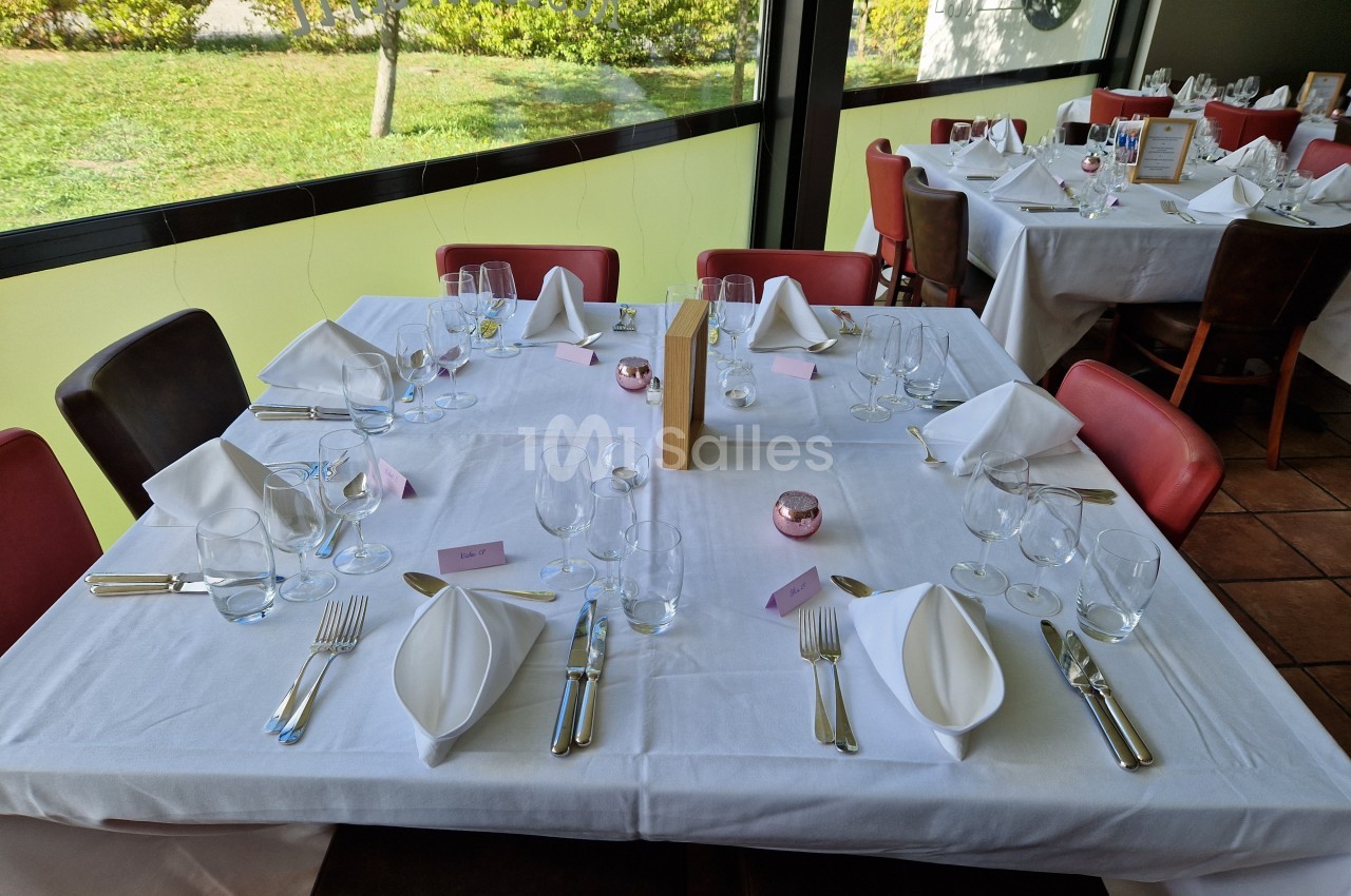 Table de restaurant dressée avec nappes blanches, serviettes pliées, verres, couverts et menus, près d'une baie vitrée.