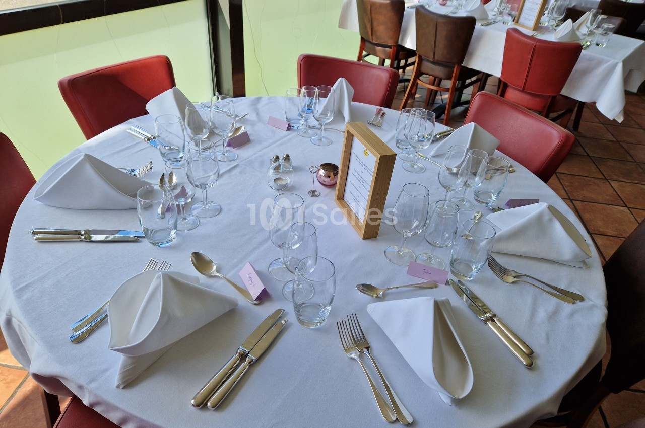 Table ronde dressée avec nappes blanches, serviettes pliées, verres et couverts, dans une salle de restaurant lumineuse.
