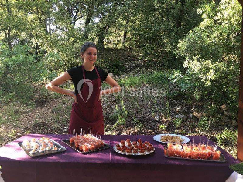 Une femme en tablier rouge présente un buffet de verrines et amuse-bouches sur une table en extérieur boisé.