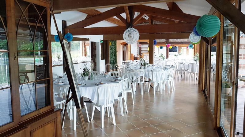 Salle de réception lumineuse avec tables rondes dressées, chaises blanches et décorations suspendues colorées.