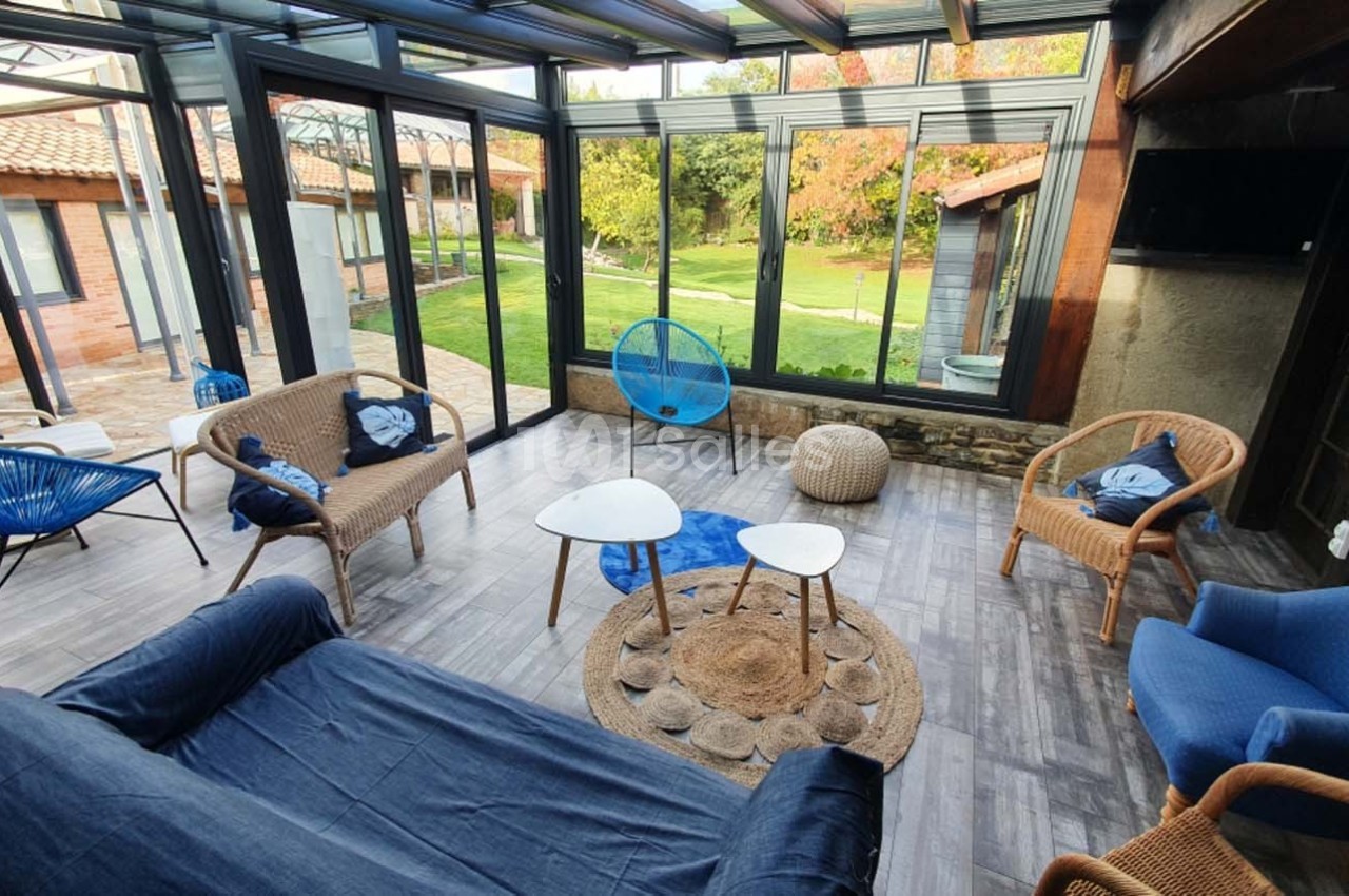 Salon lumineux avec mobilier en rotin et vue sur un jardin verdoyant à travers de grandes baies vitrées.