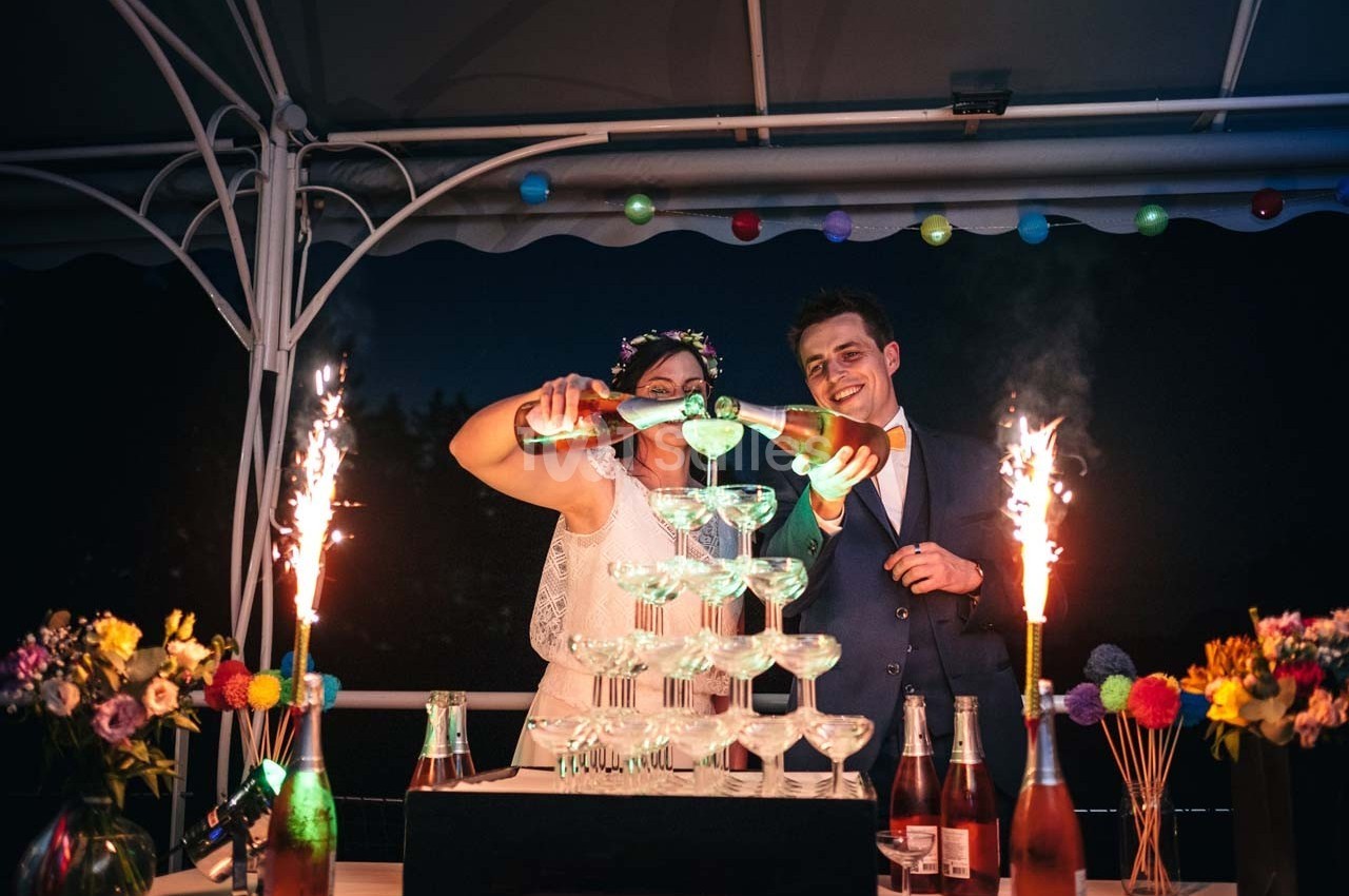 Un couple remplit une pyramide de coupes avec du champagne, entouré de décorations festives et de feux d'artifice.