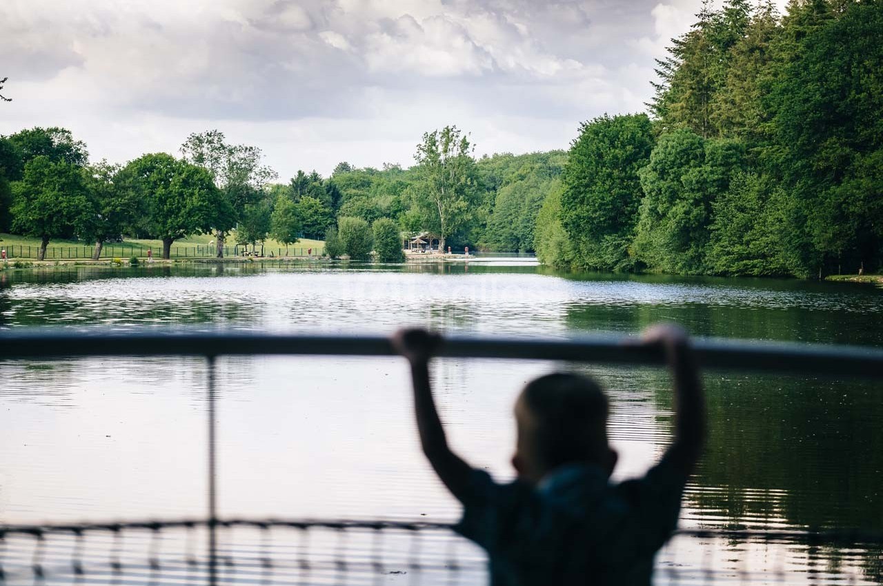 Un enfant observe un lac entouré d'arbres, sous un ciel partiellement nuageux.