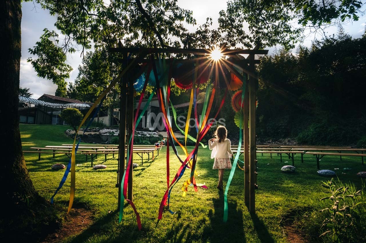 Une enfant marche sous une arche décorée de rubans colorés dans un jardin ensoleillé avec des bancs en arrière-plan.