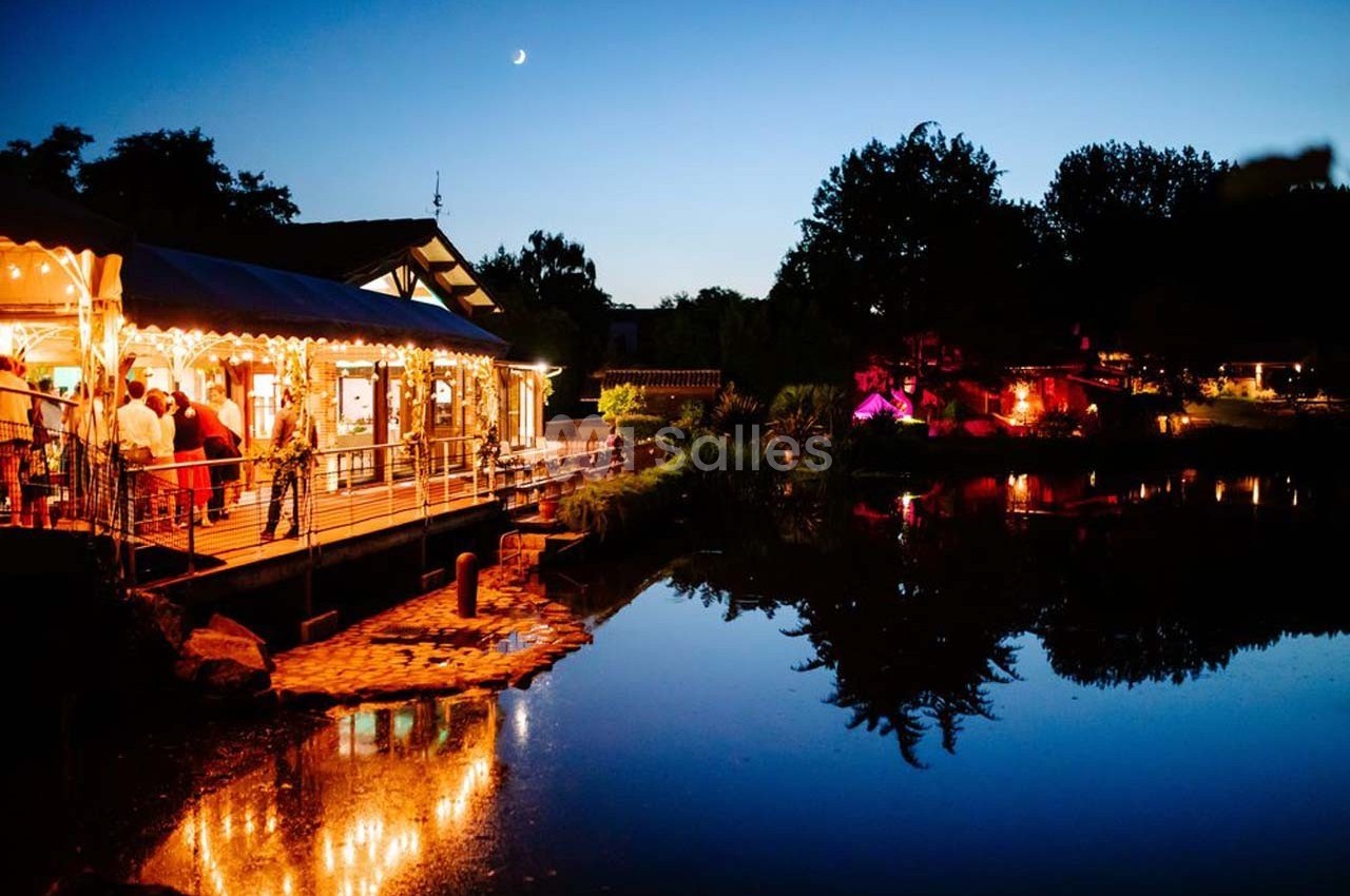 Restaurant éclairé en soirée au bord d'un étang calme, avec reflets des lumières sur l'eau et ciel crépusculaire.