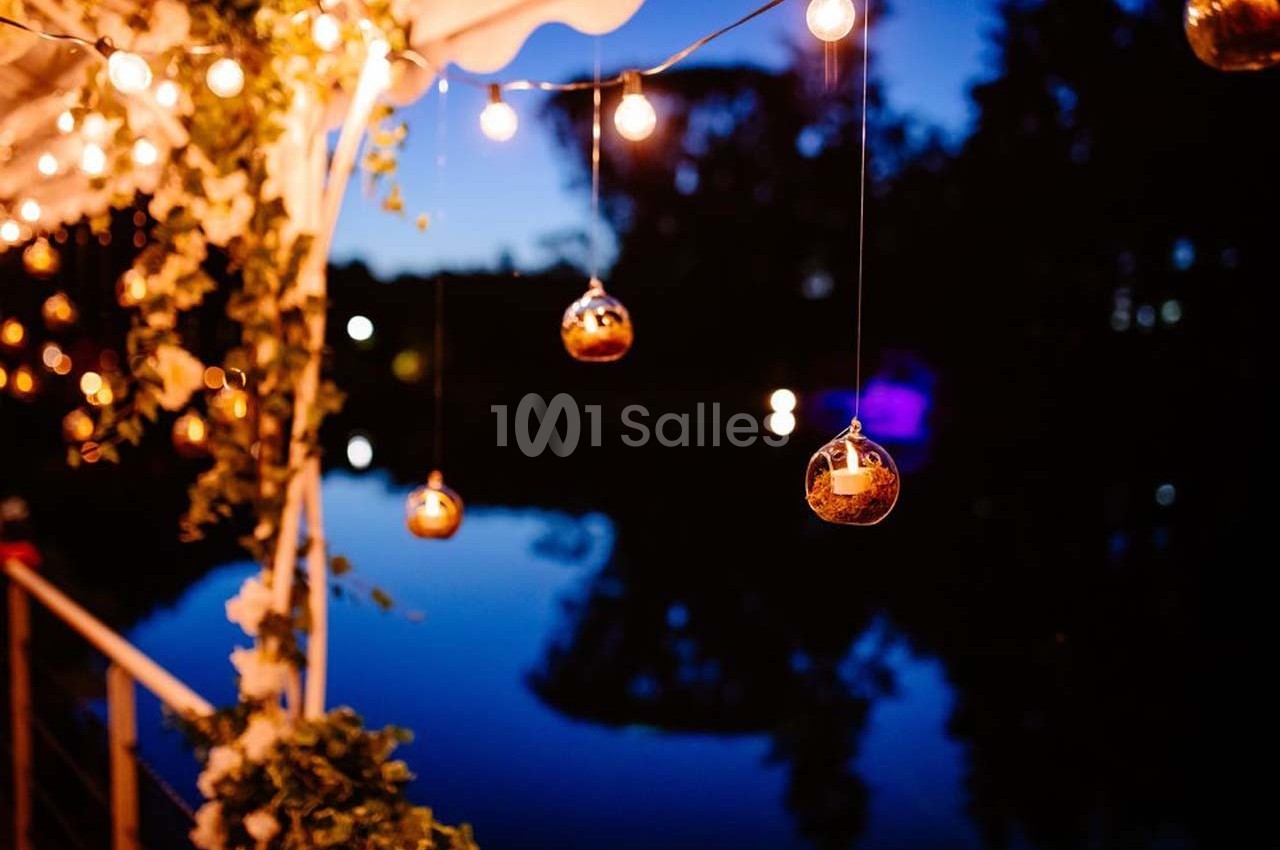 Guirlandes lumineuses et décorations suspendues éclairent un espace extérieur près d'un plan d'eau au crépuscule.