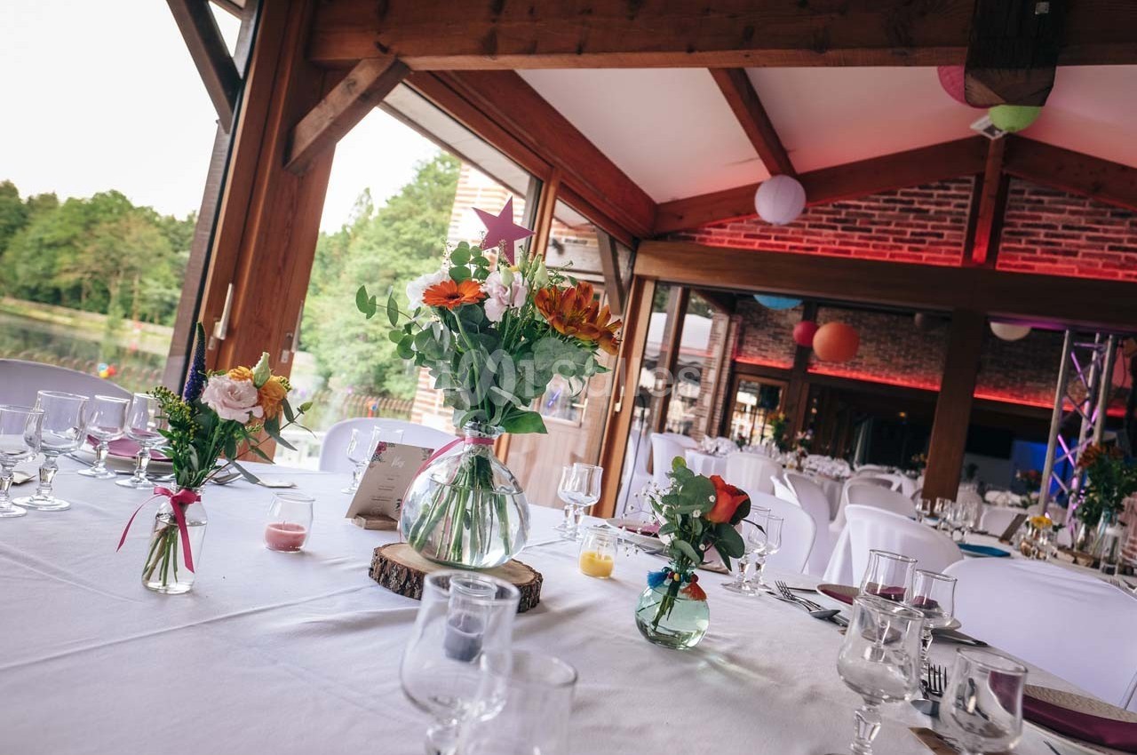 Salle de réception décorée avec des tables dressées, des vases fleuris et une vue sur l'extérieur verdoyant.