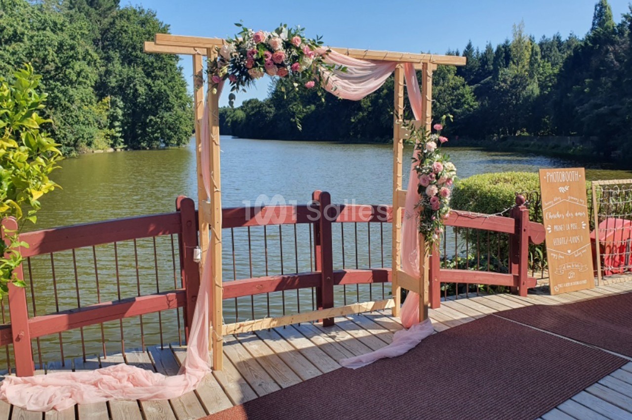 Arche en bois décorée de fleurs et de voilages roses, installée sur une terrasse en bois au bord d'un lac.