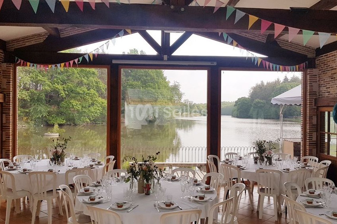 Salle de réception décorée avec des guirlandes colorées, tables dressées, vue sur un lac et des arbres à travers de grandes…