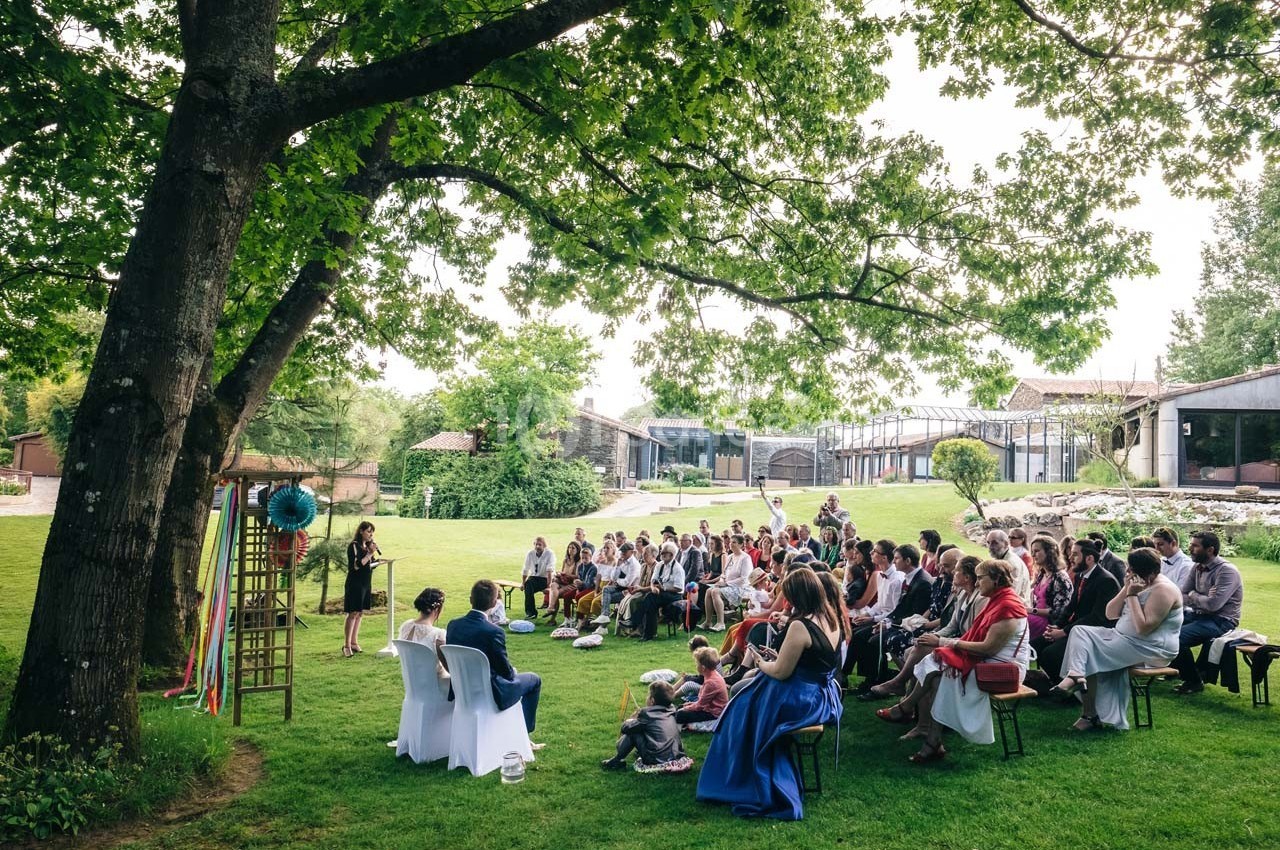 Un groupe de personnes assises en plein air sous un grand arbre, écoutant une femme debout qui s'exprime.
