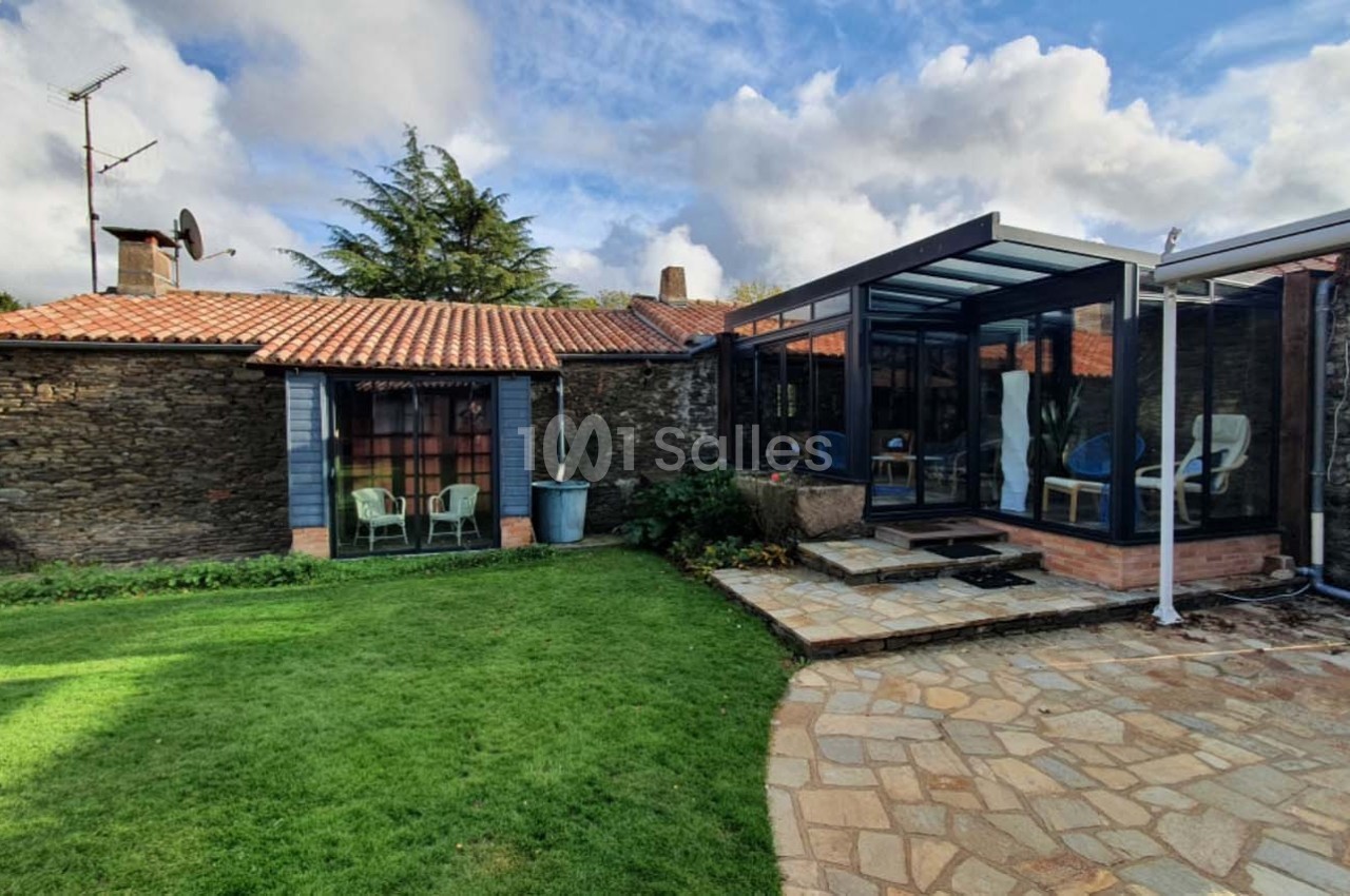 Maison en pierre avec toit en tuiles, véranda moderne en verre et jardin avec pelouse et terrasse en pierre.