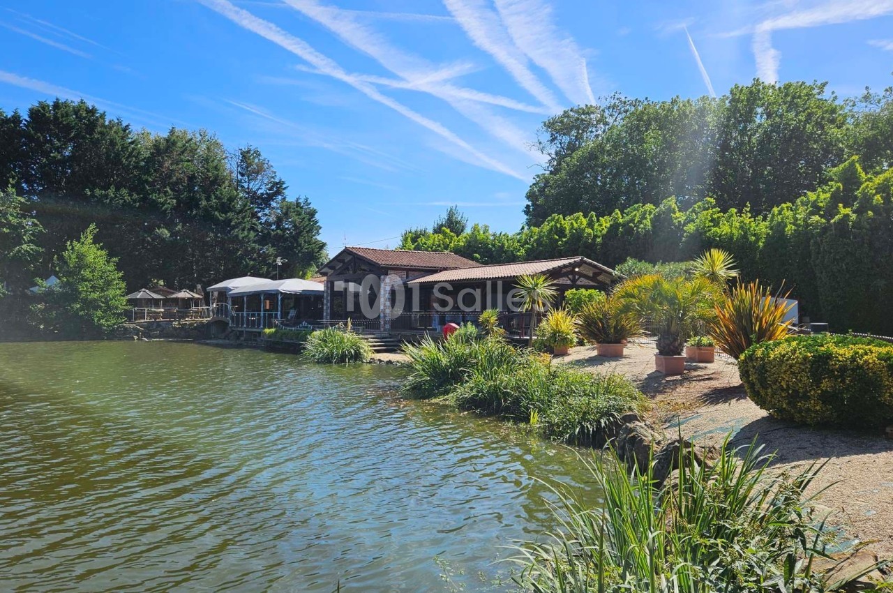 Bâtiment en bois avec terrasse au bord d'un étang, entouré de végétation et éclairé par un ciel ensoleillé.