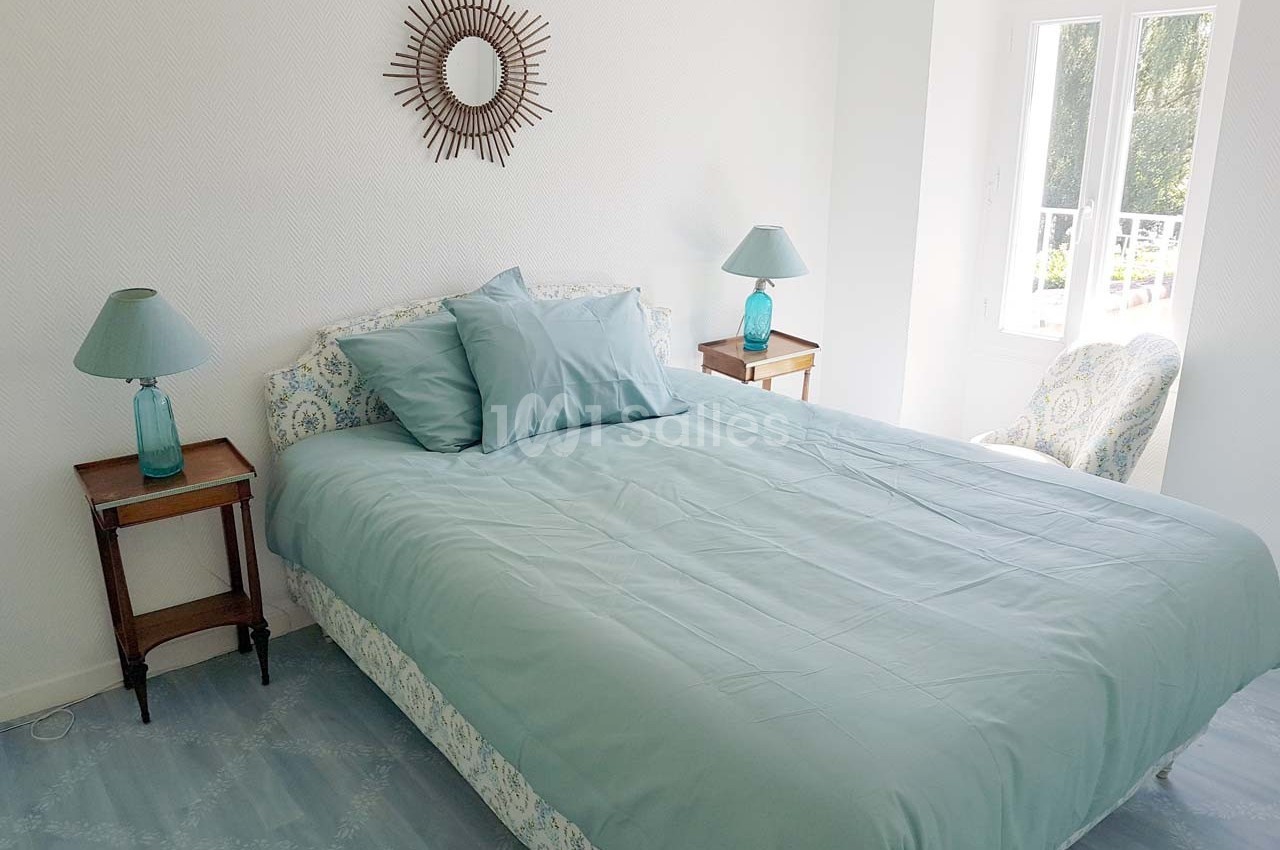 Chambre lumineuse avec un lit double, draps bleus, tables de chevet en bois et une fenêtre donnant sur l'extérieur.