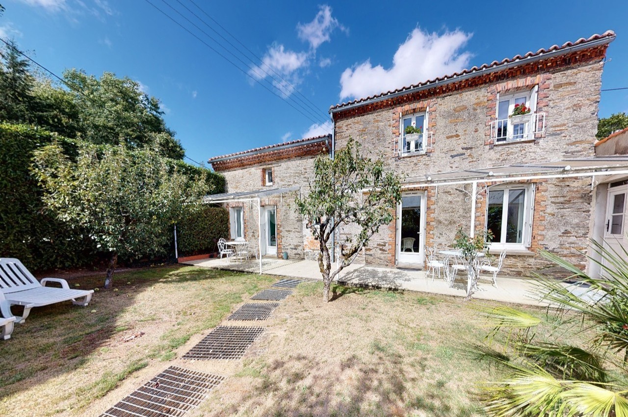 Façade en pierre d'une maison avec jardin, terrasse, chaises longues et arbres sous un ciel bleu.