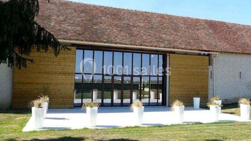 Façade d'un bâtiment avec une grande baie vitrée, entourée de murs en bois et de pots de fleurs sur une terrasse blanche.