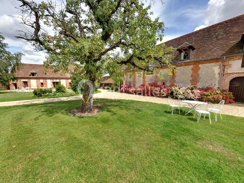 Cour verdoyante avec un arbre central, entourée de bâtiments en pierre et de fleurs colorées, sous un ciel dégagé.