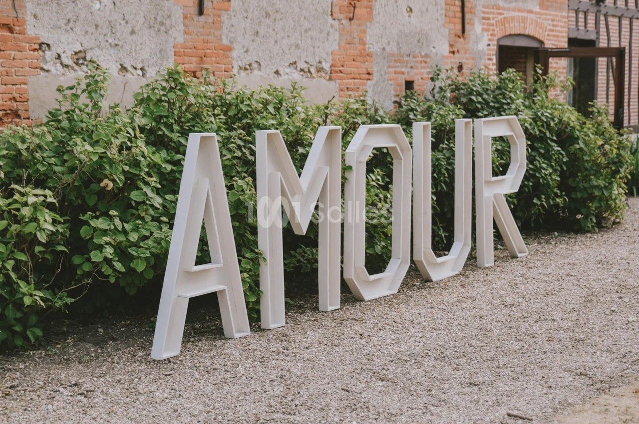 Lettres blanches formant le mot ’AMOUR’ posées devant un mur en briques et des buissons verts.
