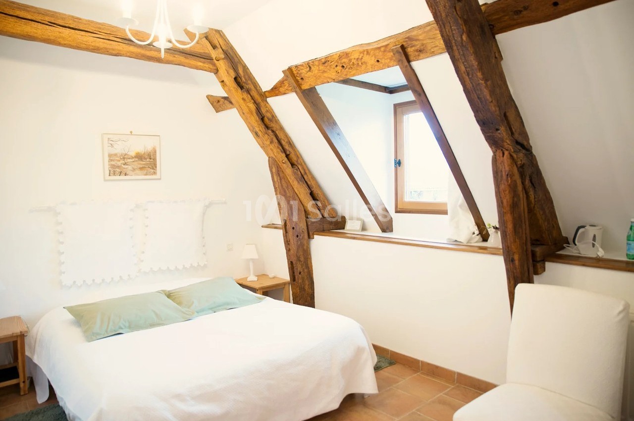 Chambre lumineuse avec poutres apparentes, lit double, murs blancs, fenêtre et décoration minimaliste.