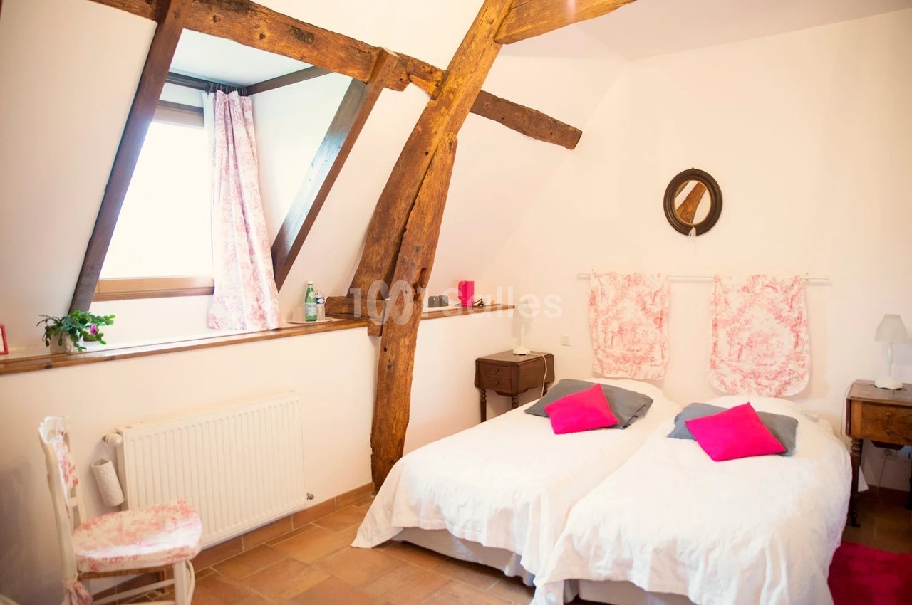 Chambre lumineuse avec poutres apparentes, deux lits simples, murs blancs et décoration rose.