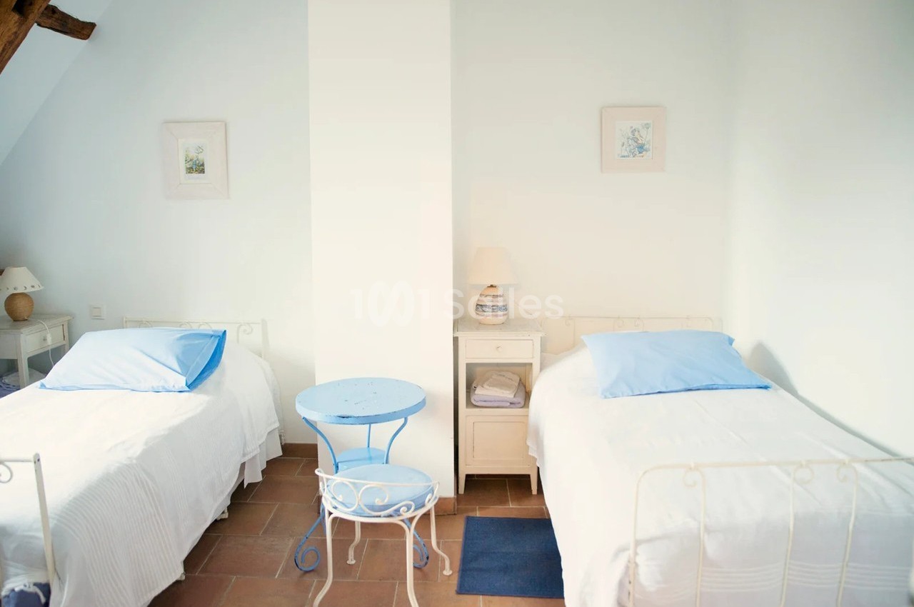 Deux lits simples avec draps blancs et oreillers bleus, une table bleue et des lampes sur des tables de chevet dans une…