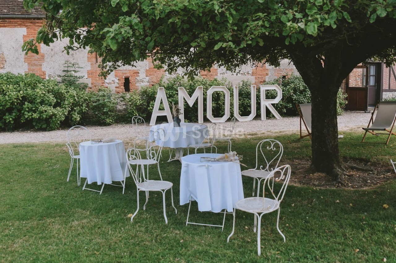 Chaises et tables blanches disposées sous un arbre, avec le mot ’AMOUR’ en grandes lettres en arrière-plan.