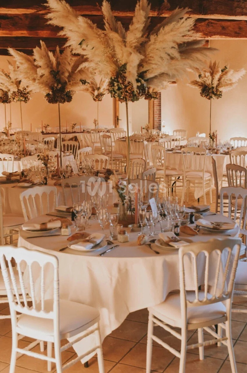 Salle de réception décorée avec des tables rondes, nappes blanches, chaises assorties et centres de table en pampas.