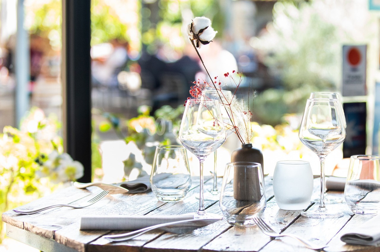 Table en bois dressée avec des verres, des couverts et une décoration florale, en terrasse lumineuse.