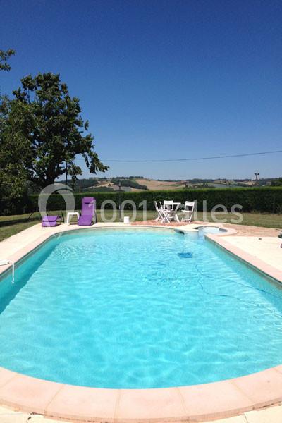 Piscine extérieure entourée de chaises longues violettes, table blanche et paysage rural en arrière-plan.