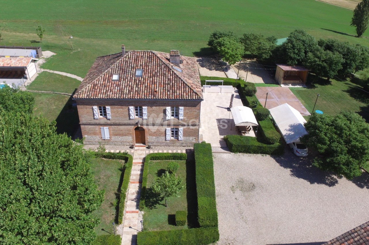 Vue aérienne d'une maison en pierre avec toit en tuiles, entourée de jardins, d'arbres et d'une cour gravillonnée.