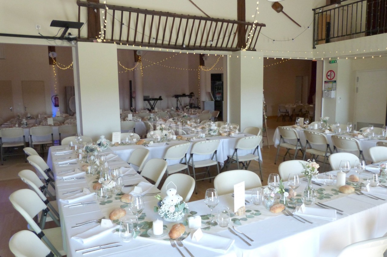 Salle de réception décorée pour un mariage, avec longues tables dressées, nappes blanches et arrangements floraux.