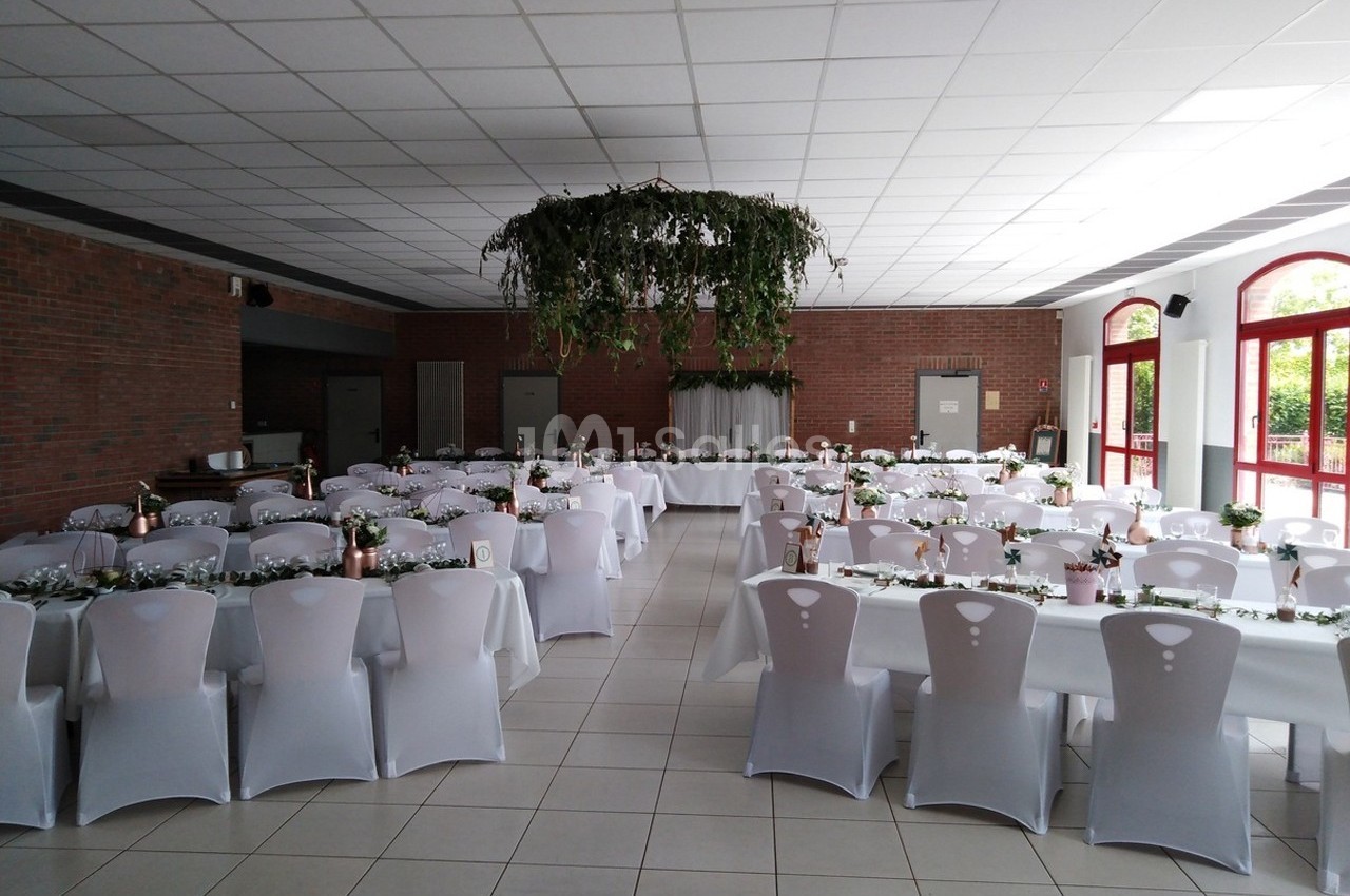 Salle de réception décorée avec des tables alignées, nappes blanches, chaises houssées et centre de table végétal suspendu.