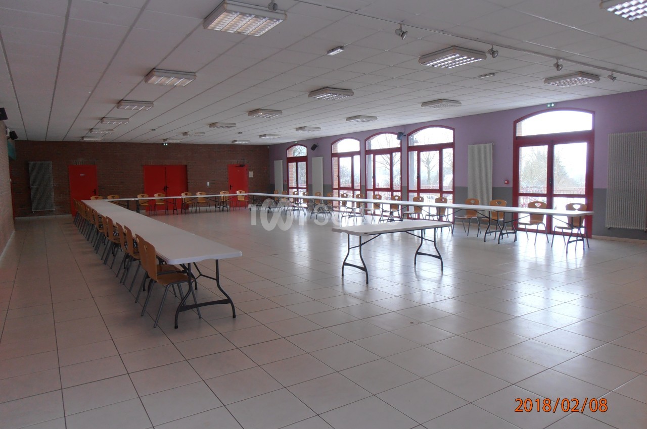 Salle spacieuse avec tables et chaises alignées, grandes fenêtres laissant entrer la lumière naturelle.