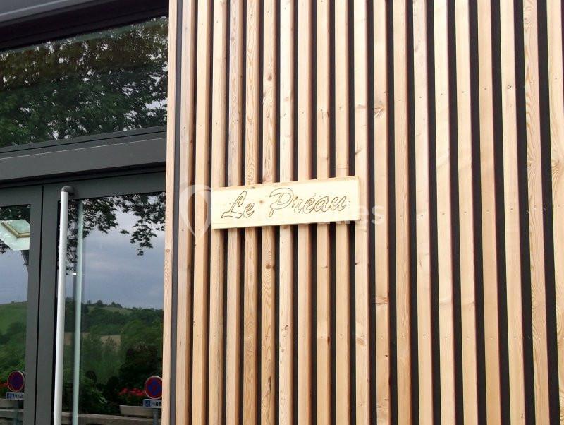 Façade en bois avec une plaque dorée portant l'inscription ’Le Préau’, près d'une porte vitrée et d'un paysage verdoyant.